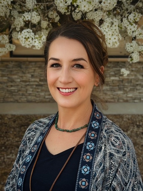 A woman with brown hair in an updo, wearing a patterned blue cardigan, black top, and green beaded necklace, smiles outdoors in front of a stone wall and blooming white flowers.