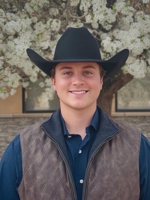 A young man wearing a black cowboy hat, blue shirt, and brown quilted vest stands smiling in front of a flowering tree with white blossoms.