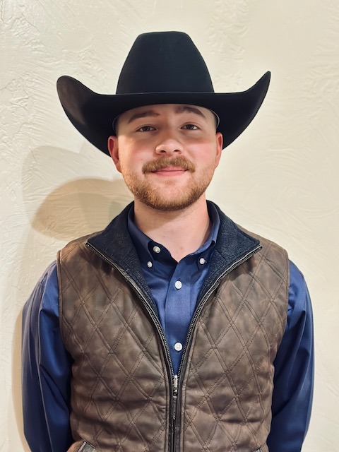 A young man with a trimmed beard wearing a black cowboy hat, navy blue shirt, and brown quilted vest stands in front of a light-textured wall, smiling slightly at the camera.
