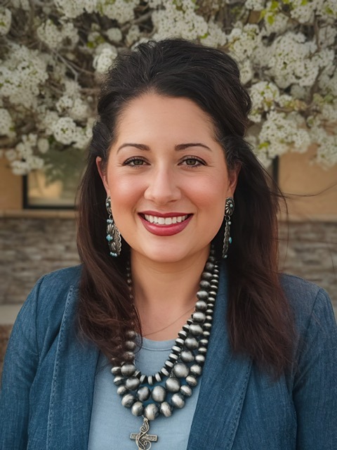 A woman with dark hair wearing a blue jacket, large silver beaded necklace, and matching earrings smiles in front of white flowering trees and a stone building.