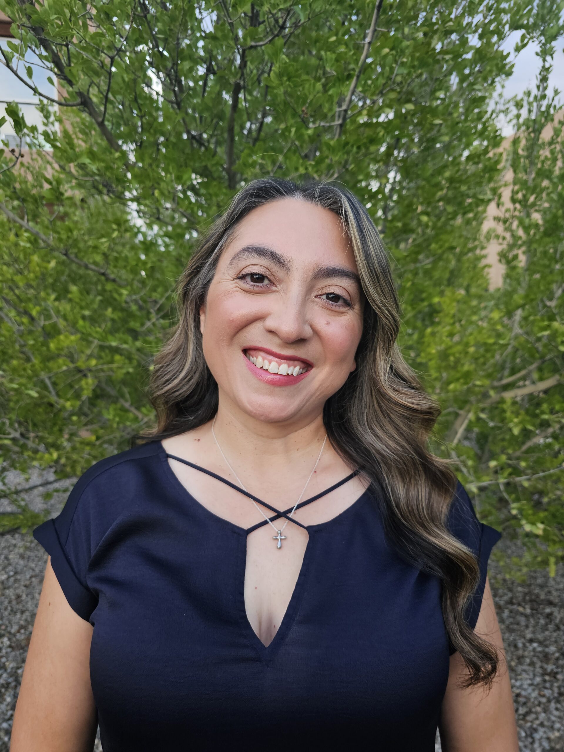 A woman with long wavy hair wearing a navy blue top and a cross necklace smiles outdoors in front of green leafy trees.