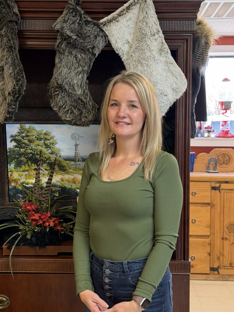A woman with straight blonde hair, wearing a long-sleeve olive green top and high-waisted jeans, stands indoors in front of a wooden cabinet decorated with faux fur stockings and a landscape painting.